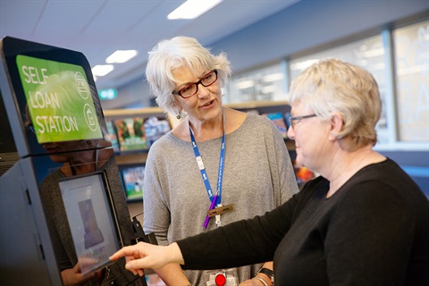 Member uses self-serve kiosk while talking to Library staff member